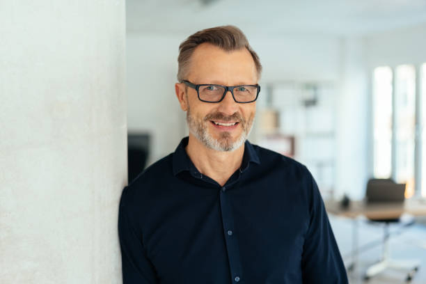 Indoor close-up portrait of a smiling handsome bearded middle-aged man in glasses and black shirt, leaning with his shoulder on white wall and looking at camera with friendly face, standing in office